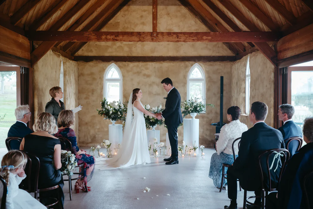Couple exchanging vows under the timber rafters of the Peppers Creek chapel, side glass doors folded open to the vineyard